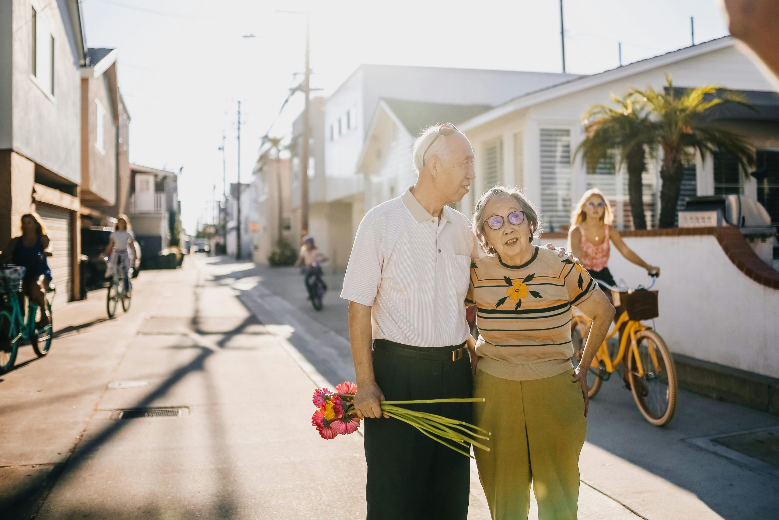 Elderly couple enjoying a walk with flowers in a sunny street setting, surrounded by cyclists.