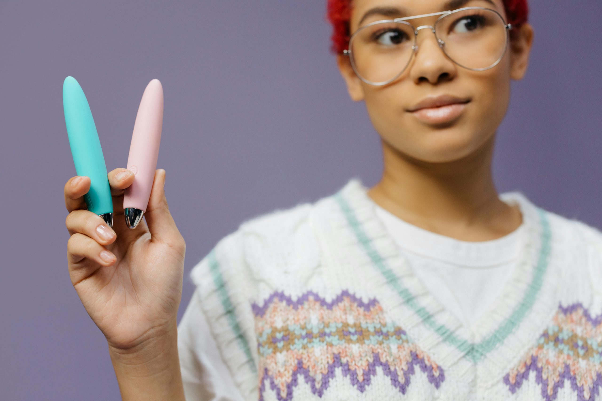 A young woman with eyeglasses holds two colorful vibrators against a purple background.