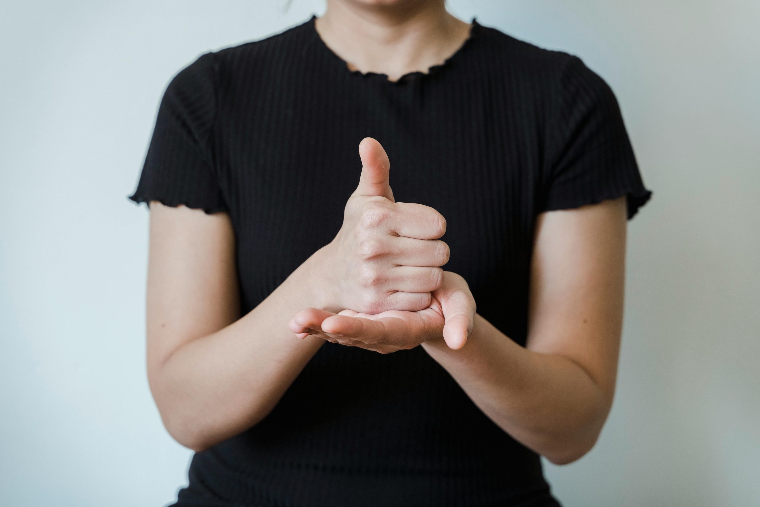 Close-up of a woman in black shirt demonstrating sign language hand gesture against plain background.