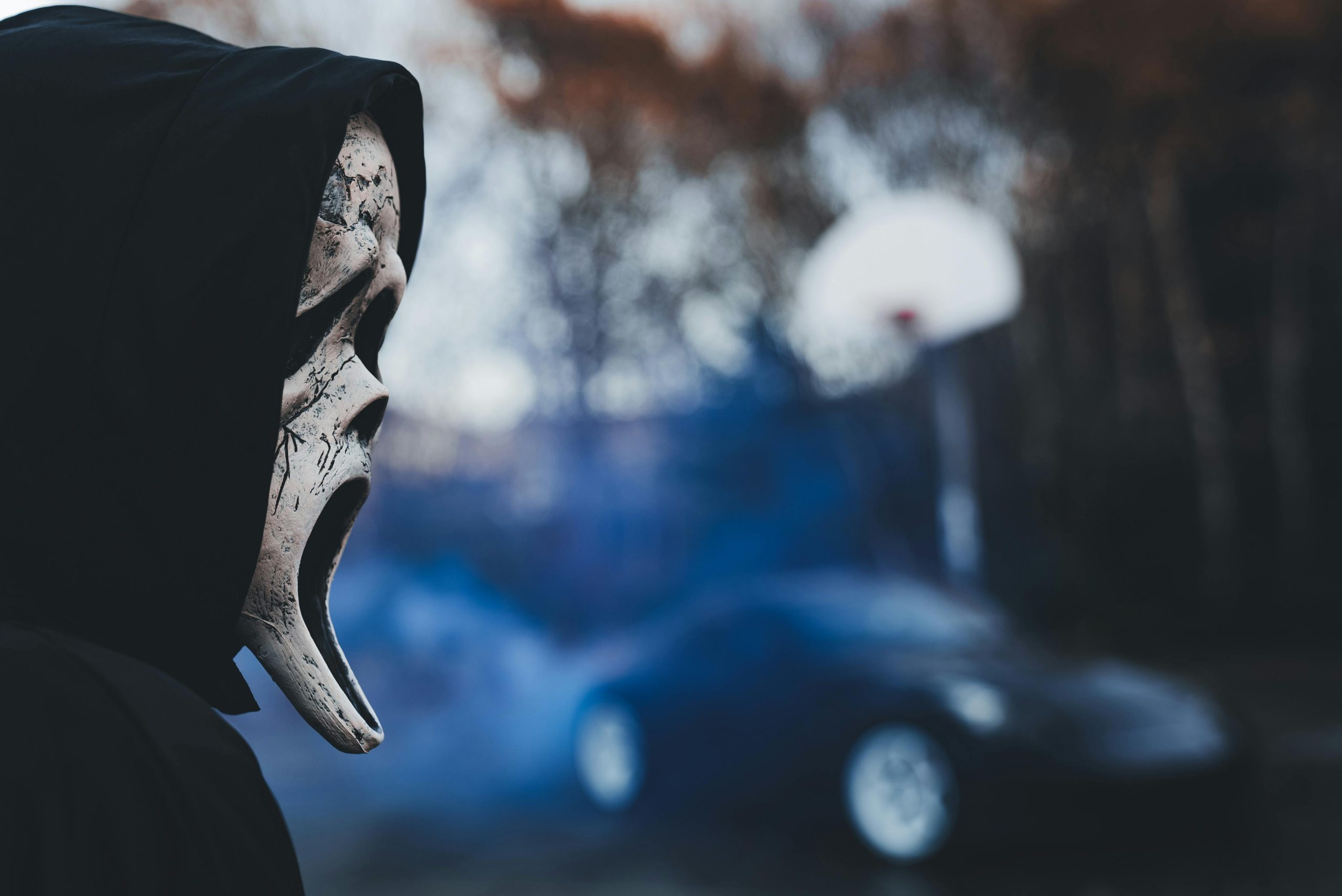 Hooded figure wearing a ghostly mask outdoors with blurred car and basketball hoop.