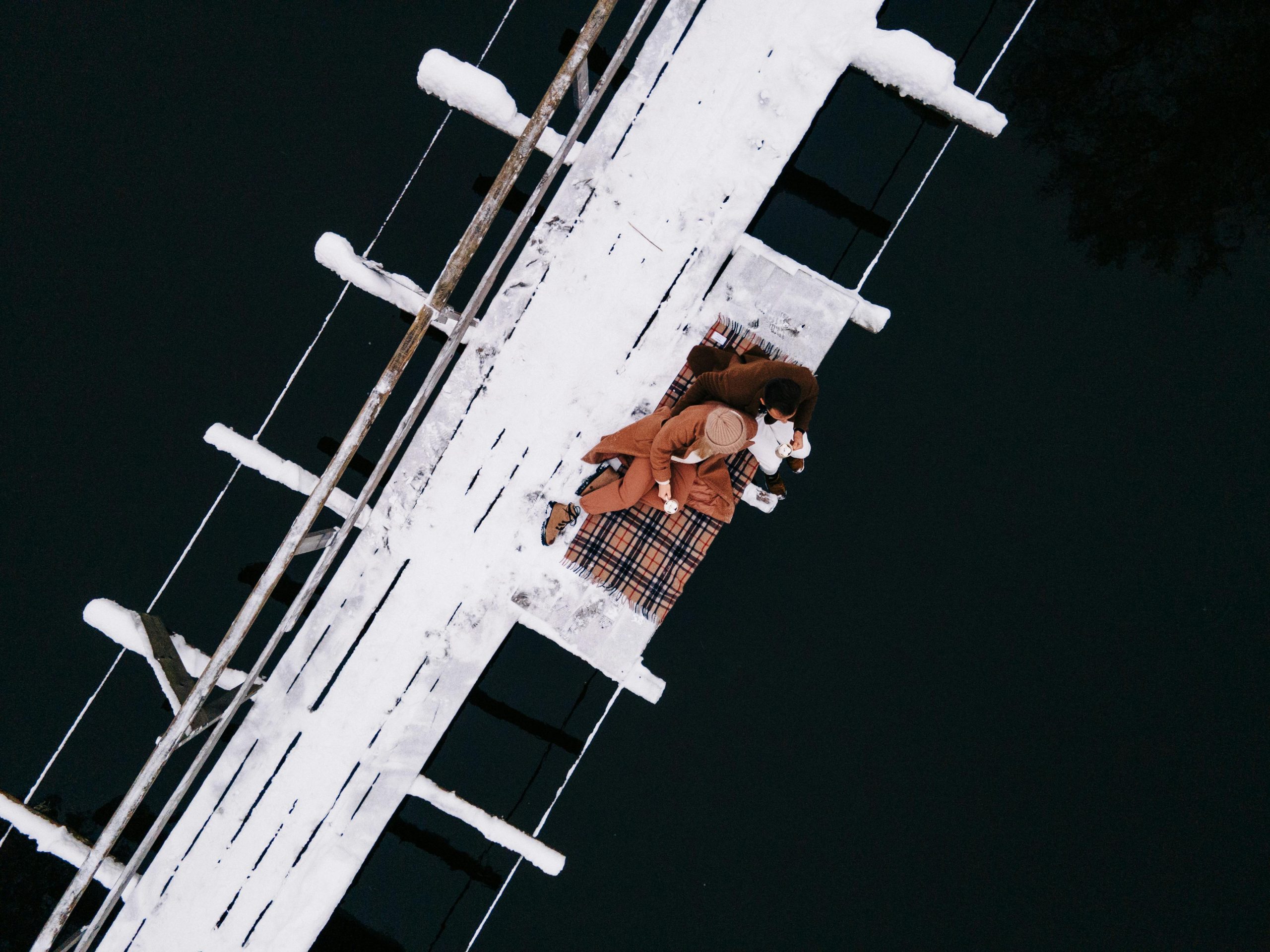 A romantic winter scene of a couple cuddling on a pier covered in snow, surrounded by water.